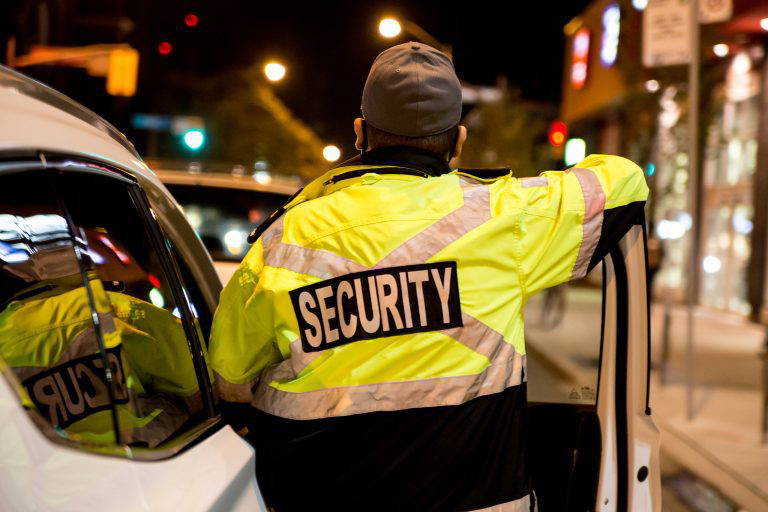 A rear view of a security guard standing next to a security car at night city.